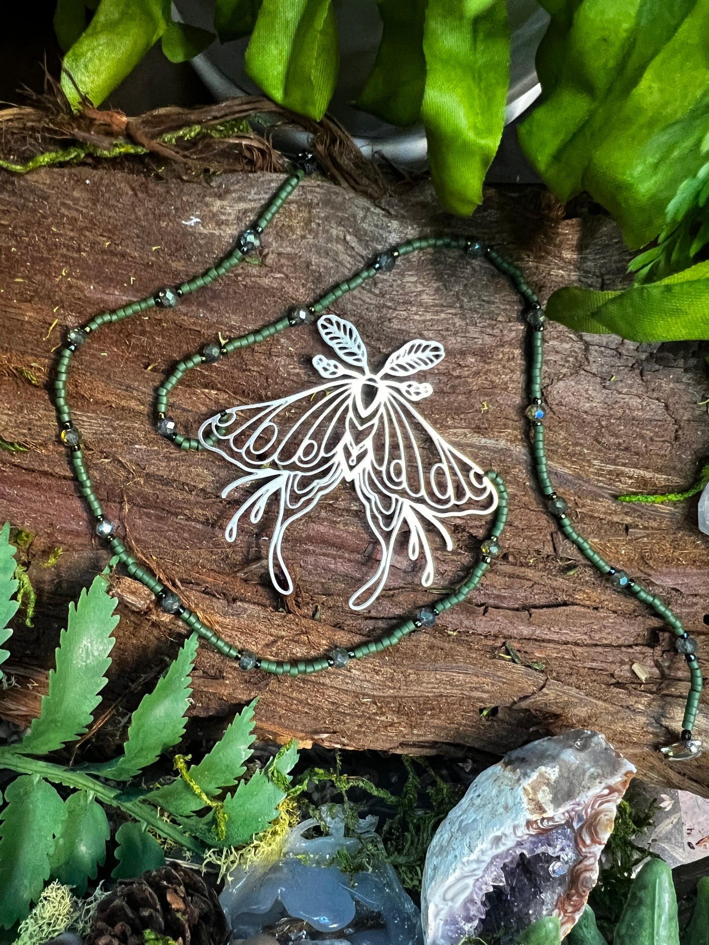 Close shot of luna moth pendant resting on moss, with forest green beads softly catching the light