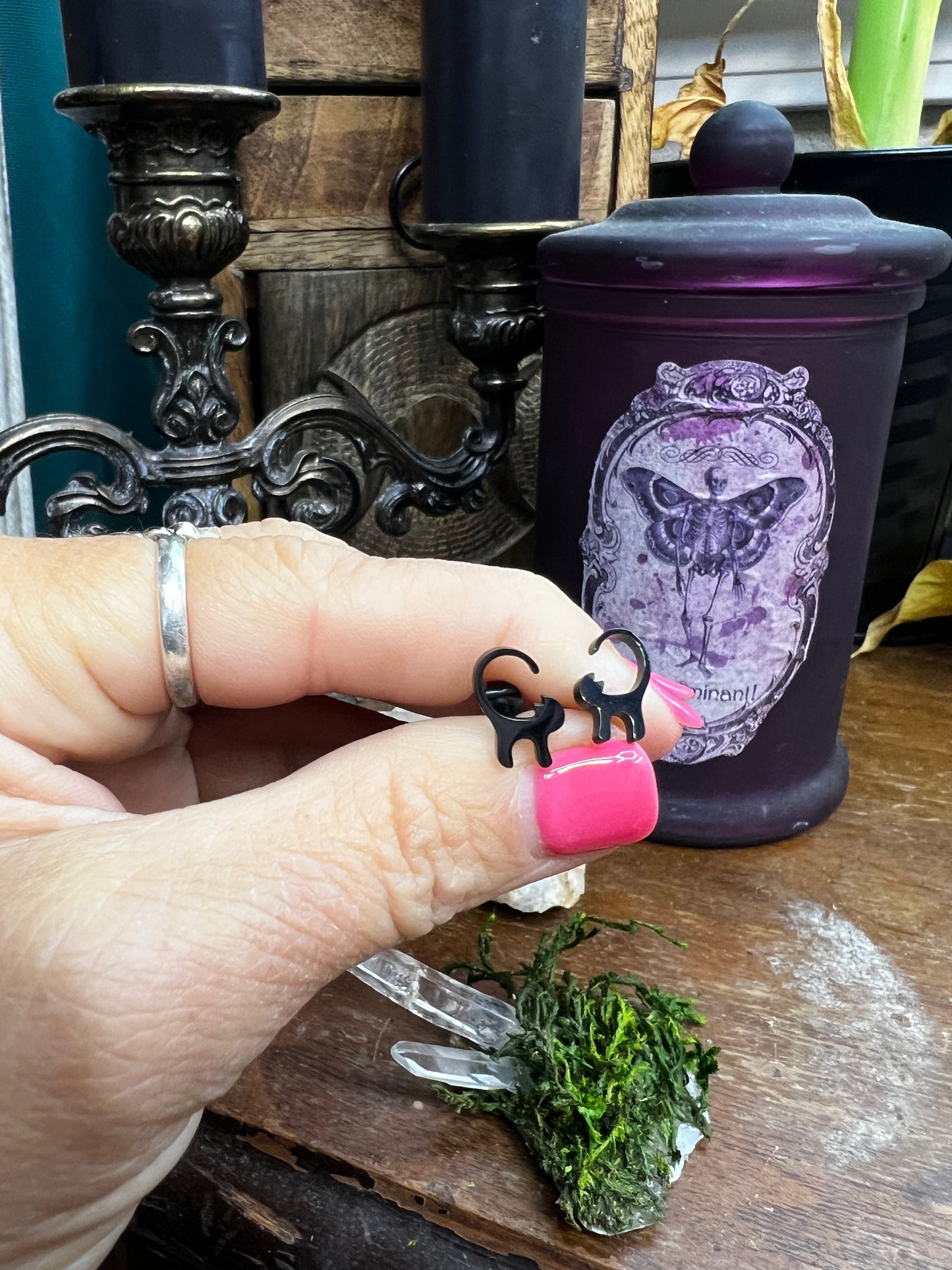 Hand holding a small black cat earrings with a purple container and candles in the background
