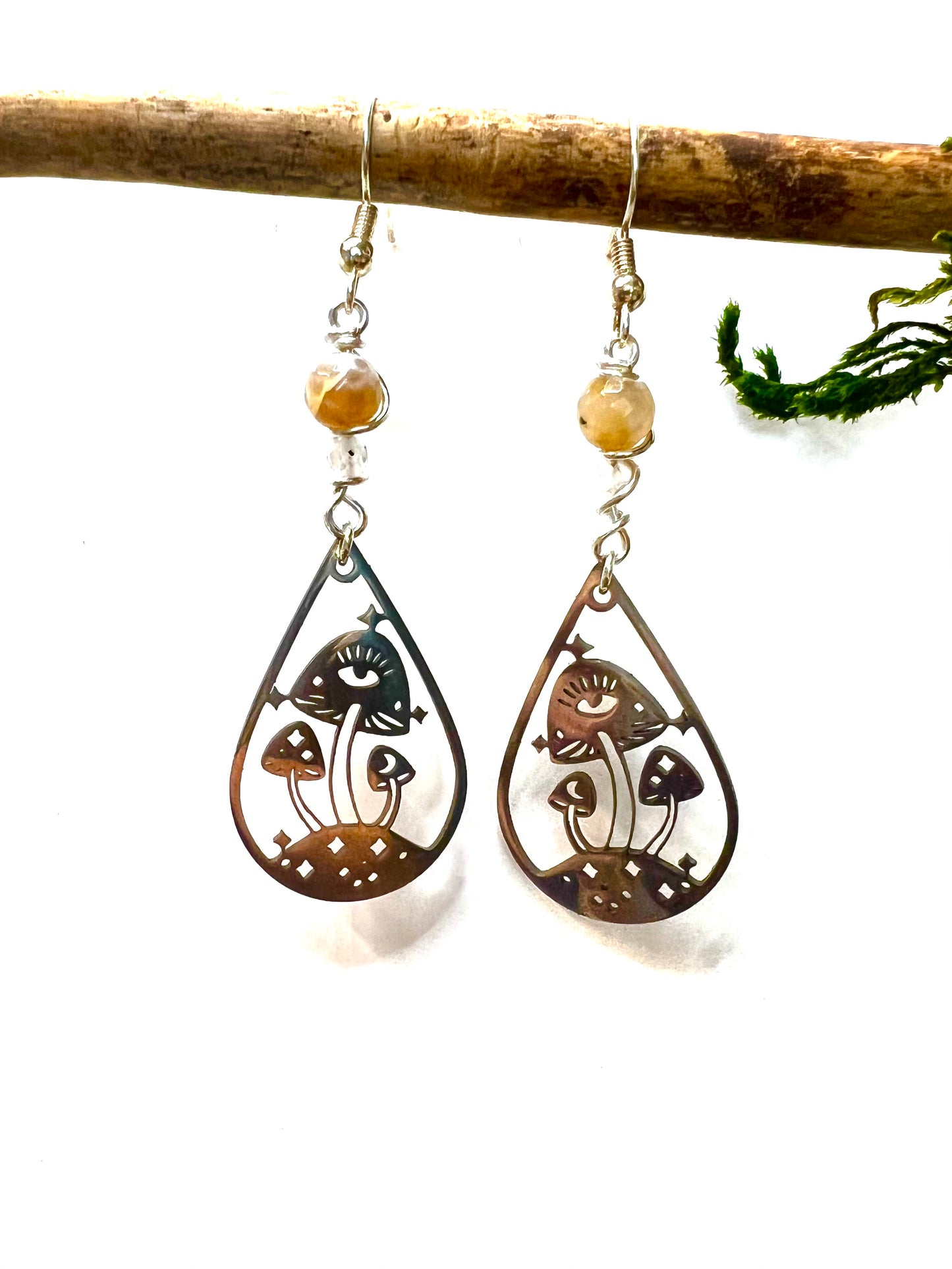 Close-up of stainless steel mushroom earrings featuring eye motifs, paired with translucent quartz and golden agate beads, hanging on hooks