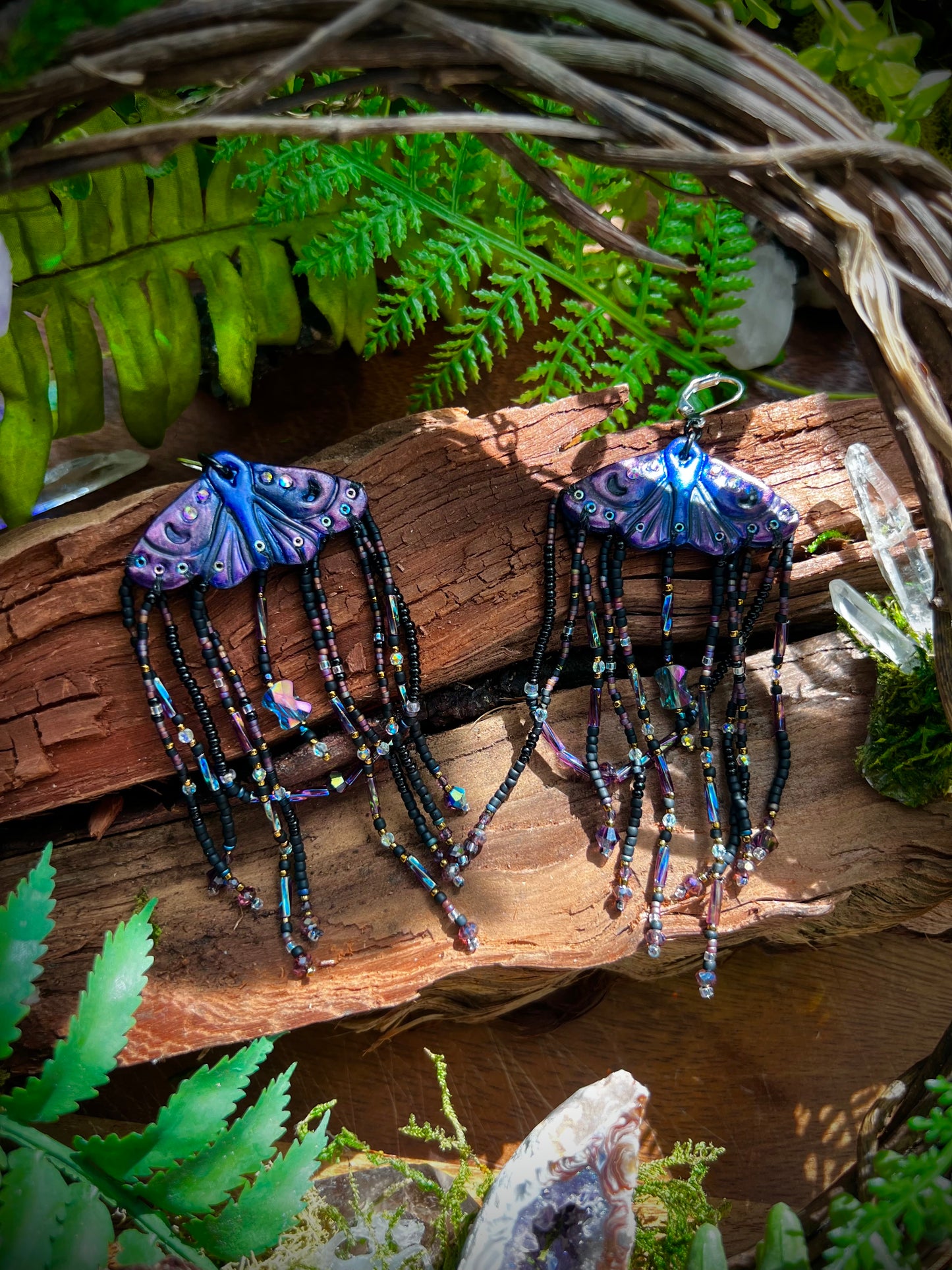 Mythical moth earrings with purple and blue polymer clay wings resting on wood with crystal details
