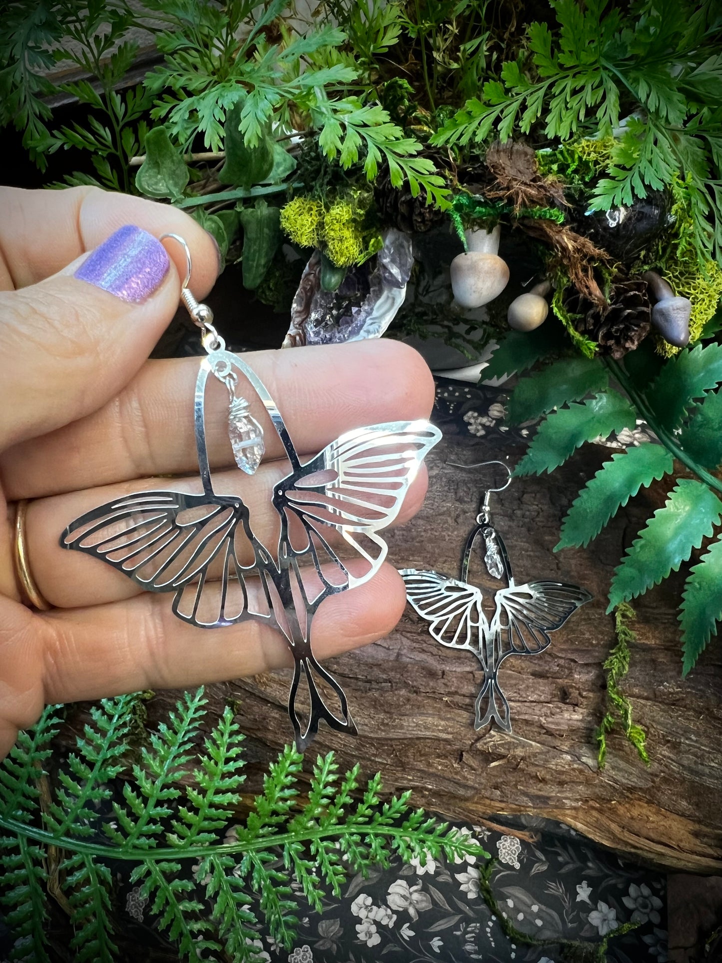 Luna moth earrings with wire-wrapped Himalayan quartz resting on bark with moss, ferns, and natural forest textures