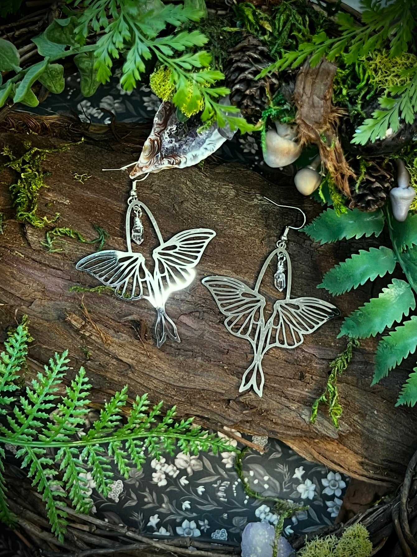Luna moth earrings with wire-wrapped Himalayan quartz resting on bark with moss, ferns, and natural forest textures