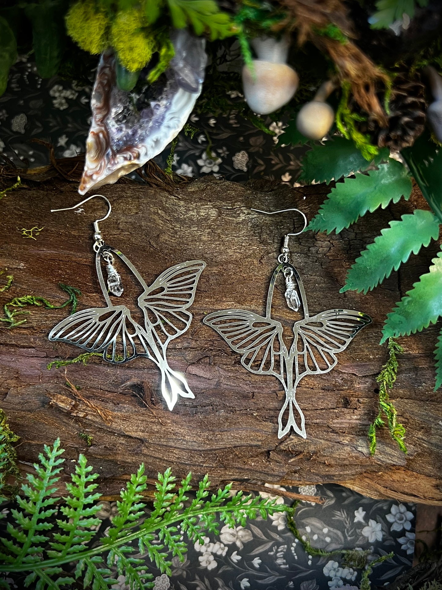 Luna moth earrings with wire-wrapped Himalayan quartz resting on bark with moss, ferns, and natural forest textures