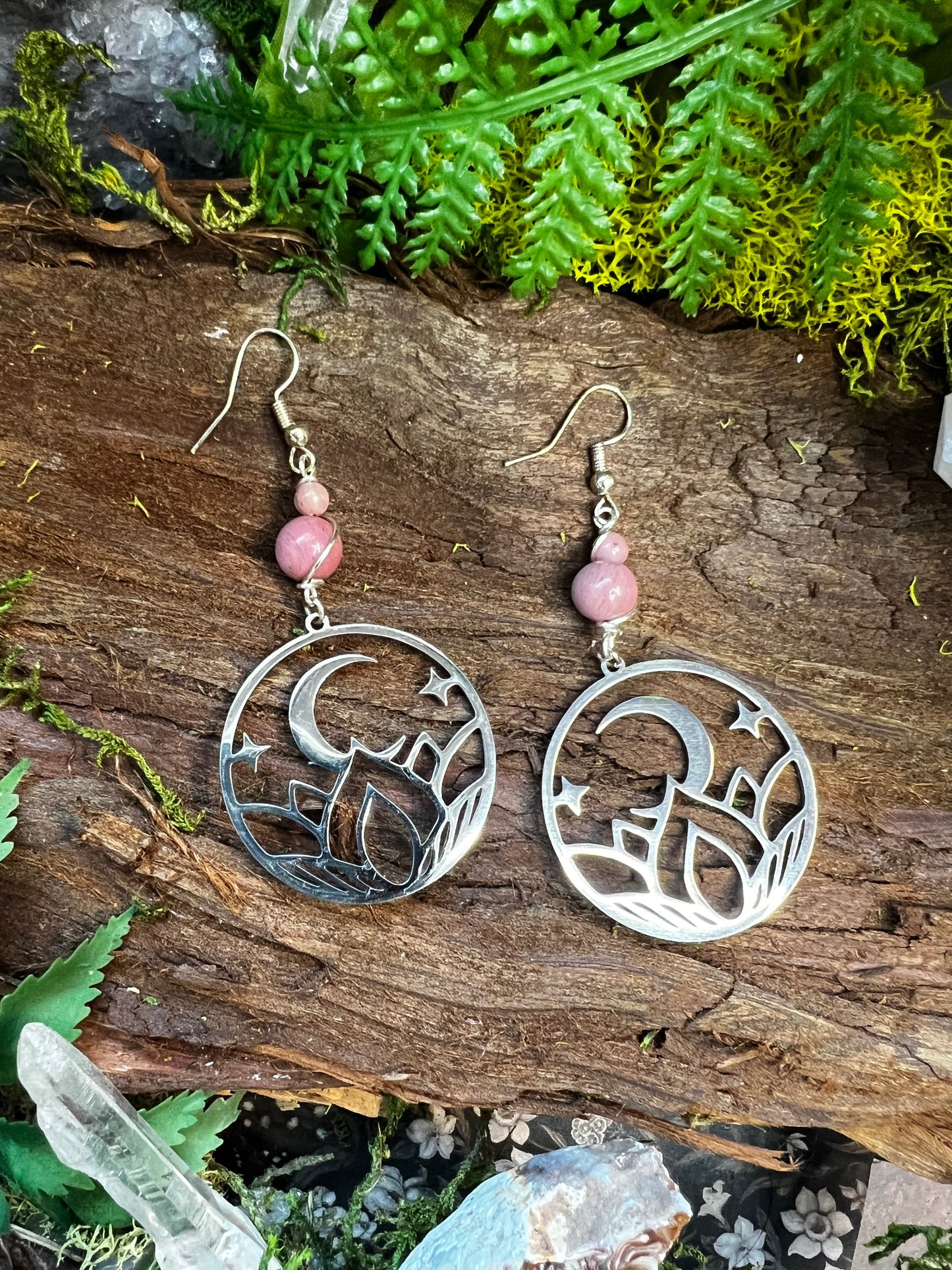 Mystical pink crystal earrings with moon and lotus charms resting on forest textures