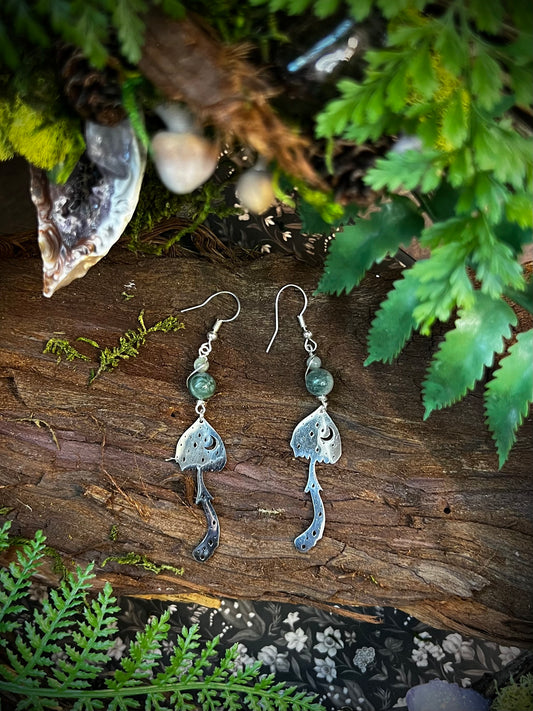 Moss agate earrings with mushroom and moon charms displayed on forest background
