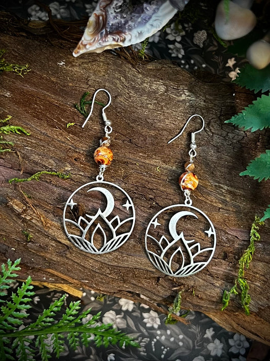 Fire opal earrings with lotus moon charms resting on a rustic wood altar surrounded by moss and crystals.