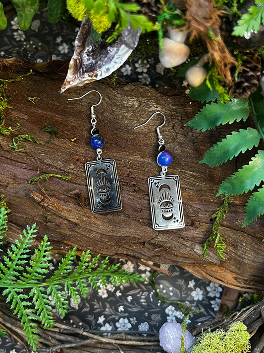 Dyed glass and iridescent tarot earrings with engraved all-seeing eye charms displayed on a wooden forest backdrop