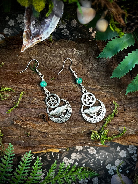 Mystical crow and pentacle earrings with green malachite beads on a wooden forest altar.