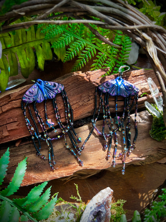Mythical moth earrings with purple and blue polymer clay wings resting on wood with crystal details