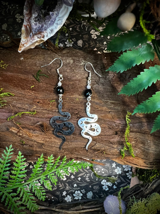 Mystical obsidian earrings with silver snake charms displayed on mossy wood