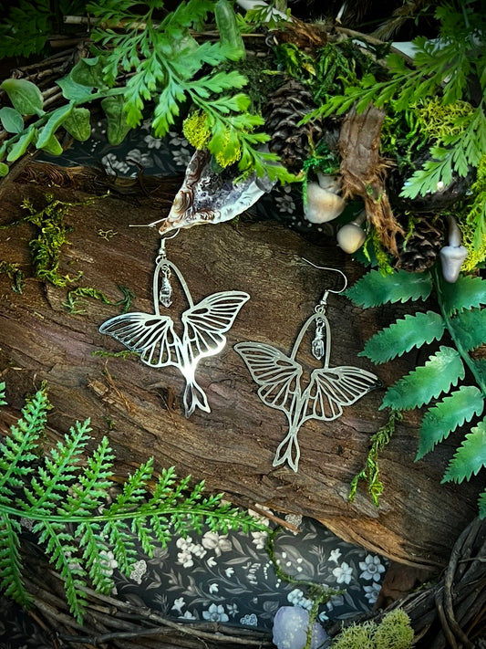 Luna moth earrings with wire-wrapped Himalayan quartz resting on bark with moss, ferns, and natural forest textures
