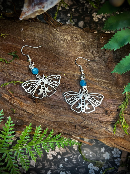 Hand holding silver moth earrings with apatite beads, surrounded by moss, ferns, and magical forest elements