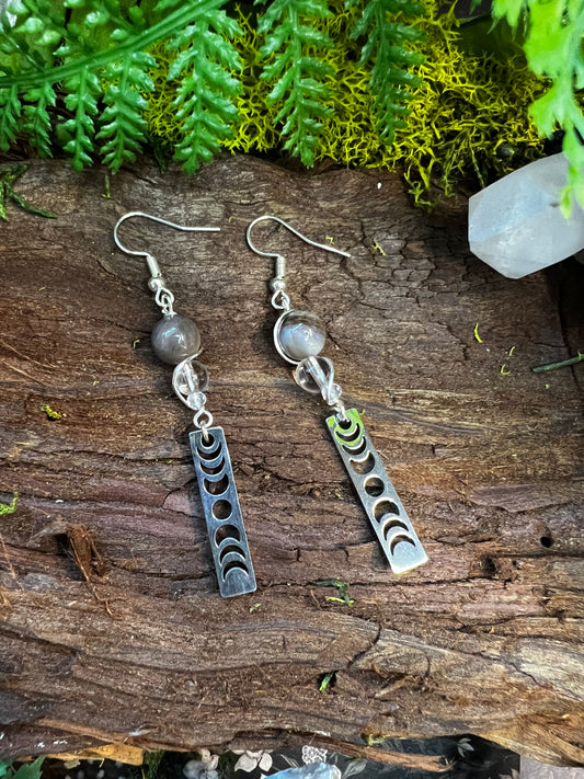 Petrified wood and clear quartz lunar phase earrings resting on mossy wood