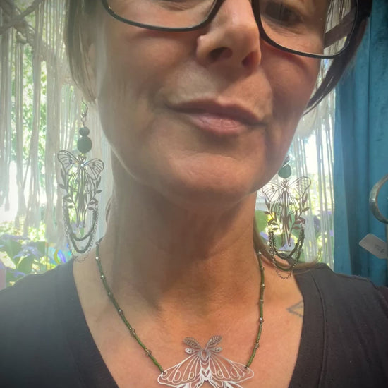 Necklace styled on mossy wood with crystals and ferns, showcasing green beaded chain and luna moth pendant