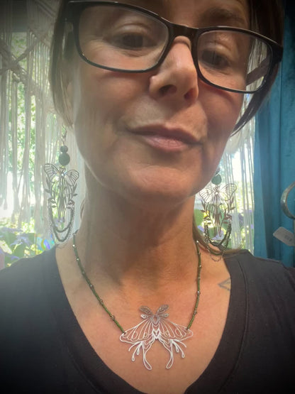 Necklace styled on mossy wood with crystals and ferns, showcasing green beaded chain and luna moth pendant