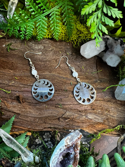 Rainbow moonstone lunar phase earrings held above wood and moss with crystals